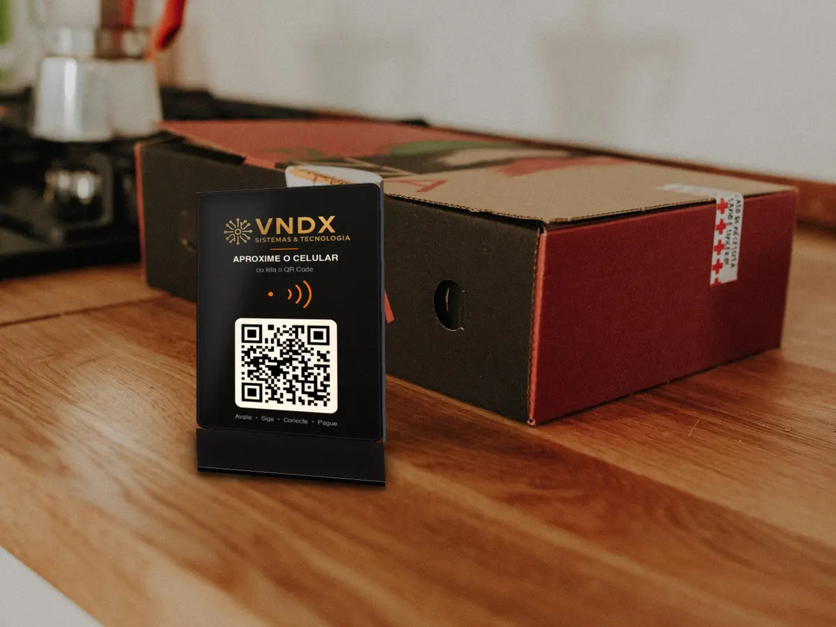 Placa VNDX Central de balcão, preta com logo, NFC e QR Code, ao lado de uma caixa de pizza no balcão de madeira