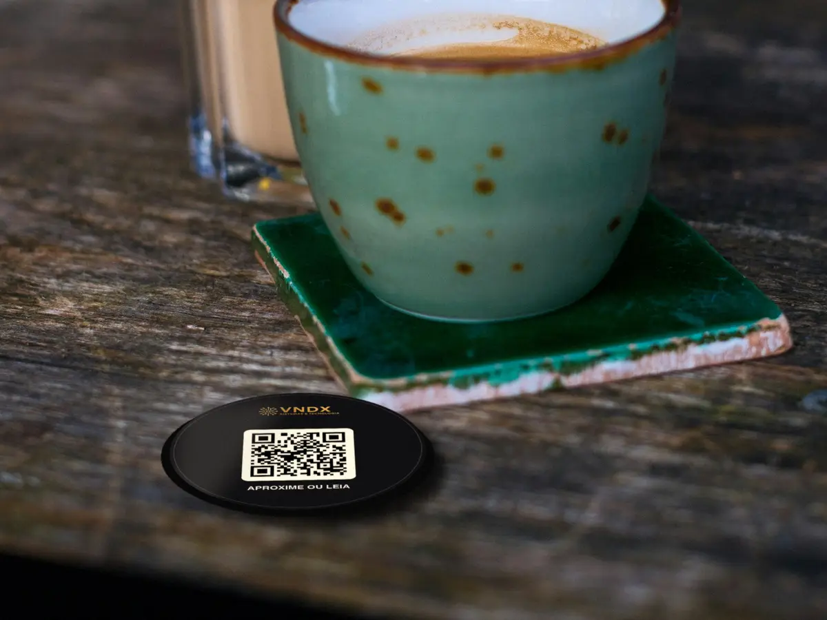 Adesivo redondo VNDX com QR Code colado na mesa de madeira de um café, ao lado do café servido